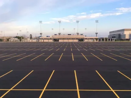 Empty asphalt parking lot in front of a stadium with bright lights under a cloudy sky.