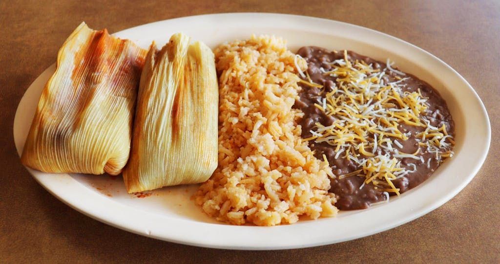A plate of food with tamales , rice and beans on a table.