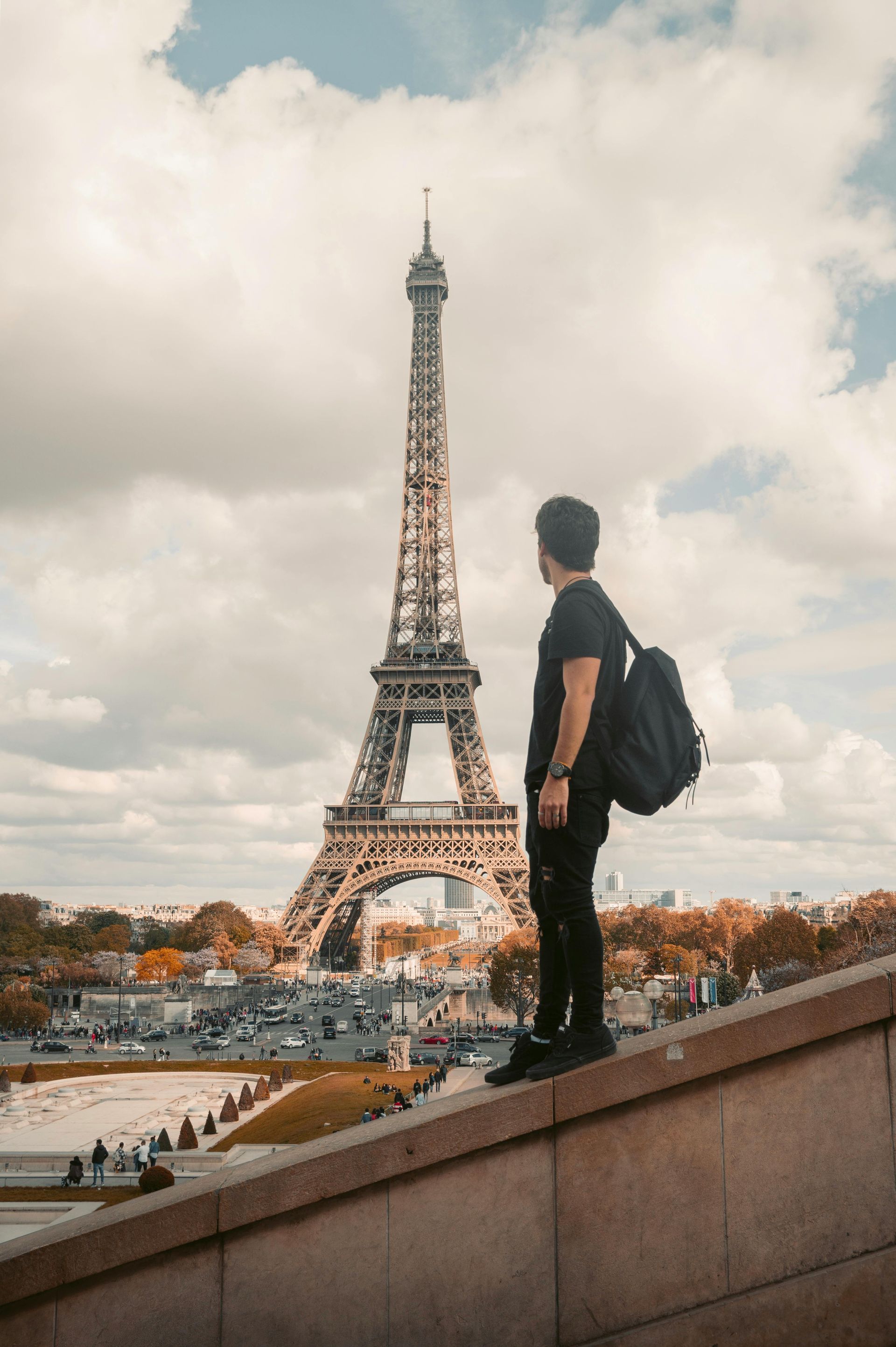 A man with a backpack is standing on a wall in front of the eiffel tower in paris.