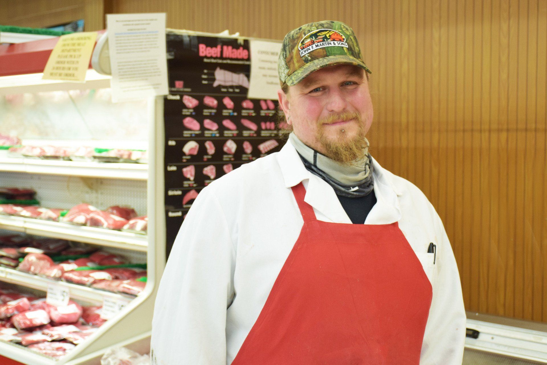 A man in a red apron is standing in front of a beef display