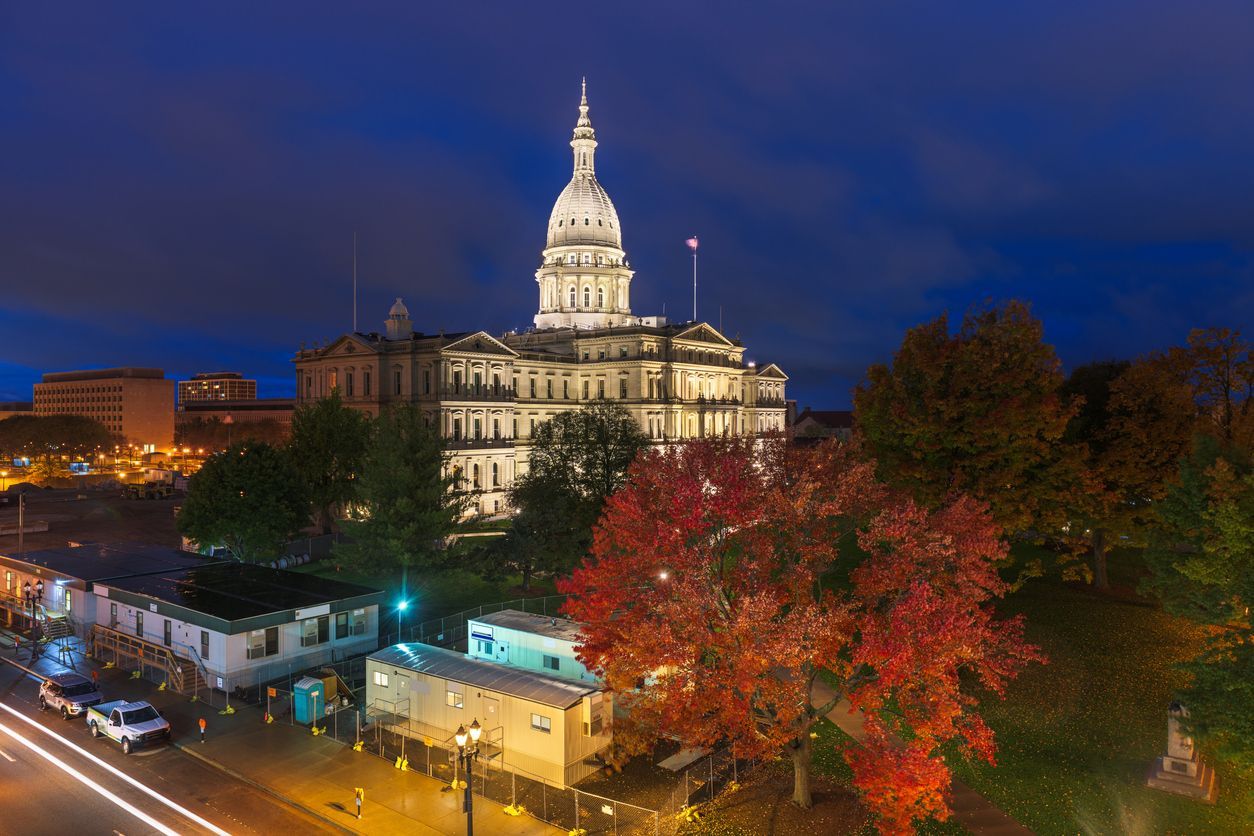 Illuminated Michigan State Capitol building at night above autumn trees and city lights