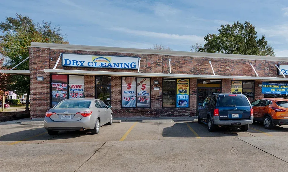Dry cleaning business storefront in a brick building with cars parked out front.