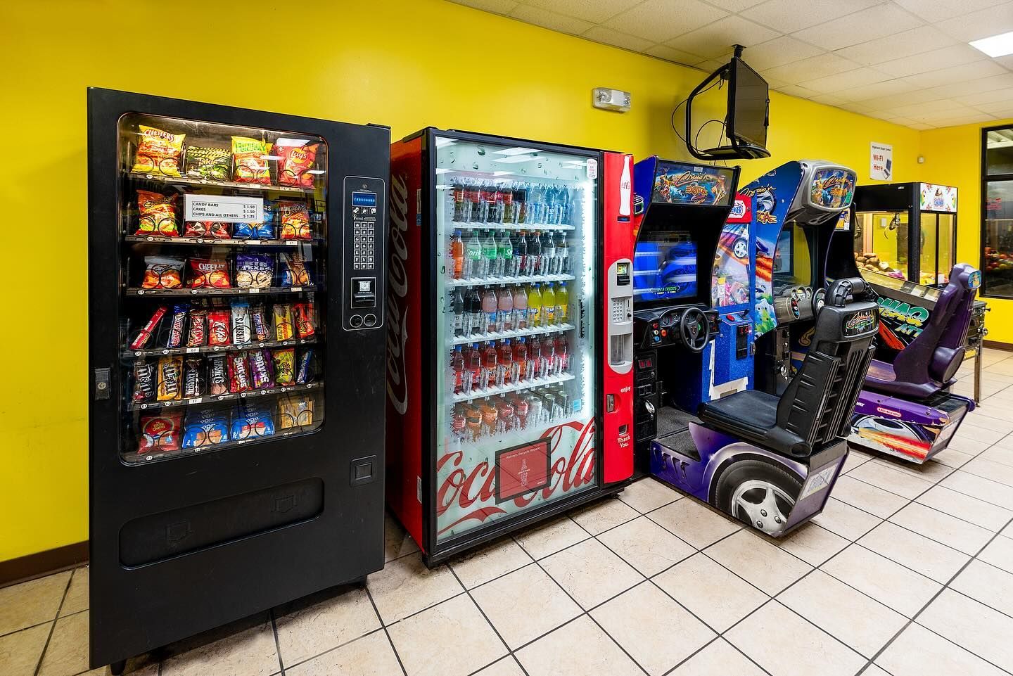 Snack and soda vending machines next to arcade games in a room with yellow walls and tile floors.