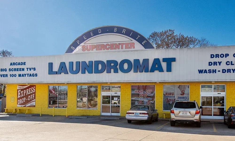 A yellow laundromat with a blue sign. A car is parked outside. The sky is bright blue.
