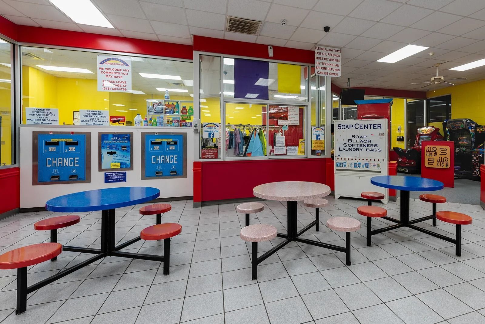 An empty laundromat interior. Red and yellow walls, coin machines, and tables with attached seats.
