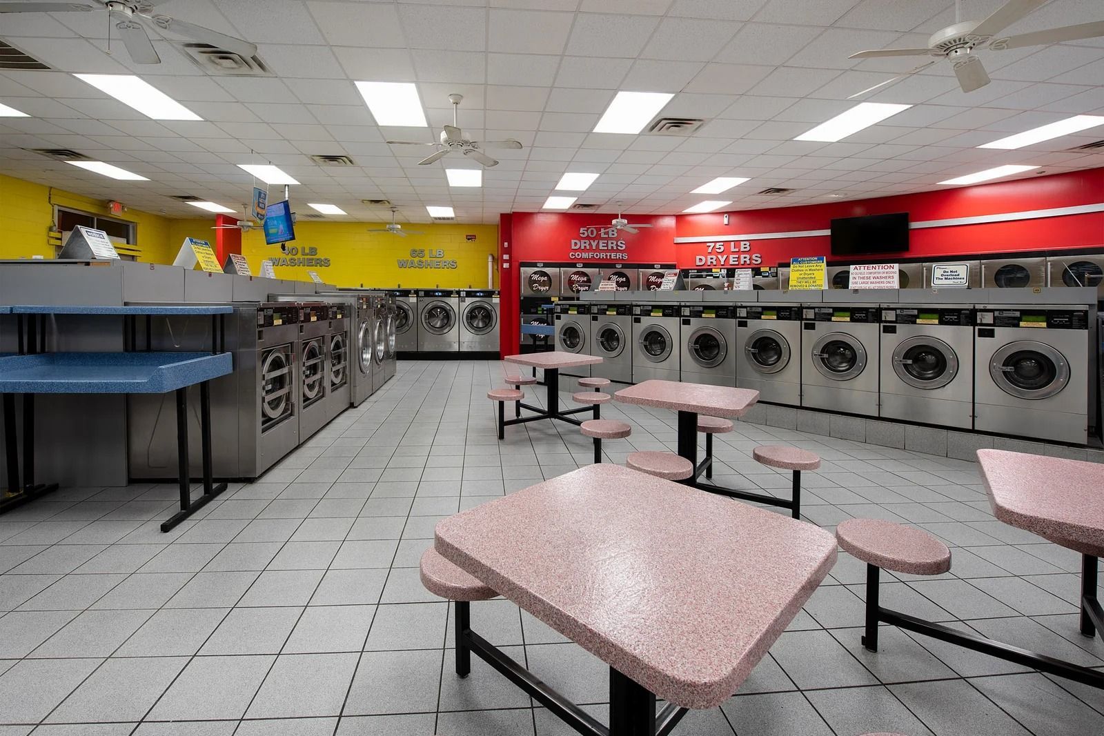 Interior of a laundromat with rows of washers and dryers, tables, and ceiling fans.