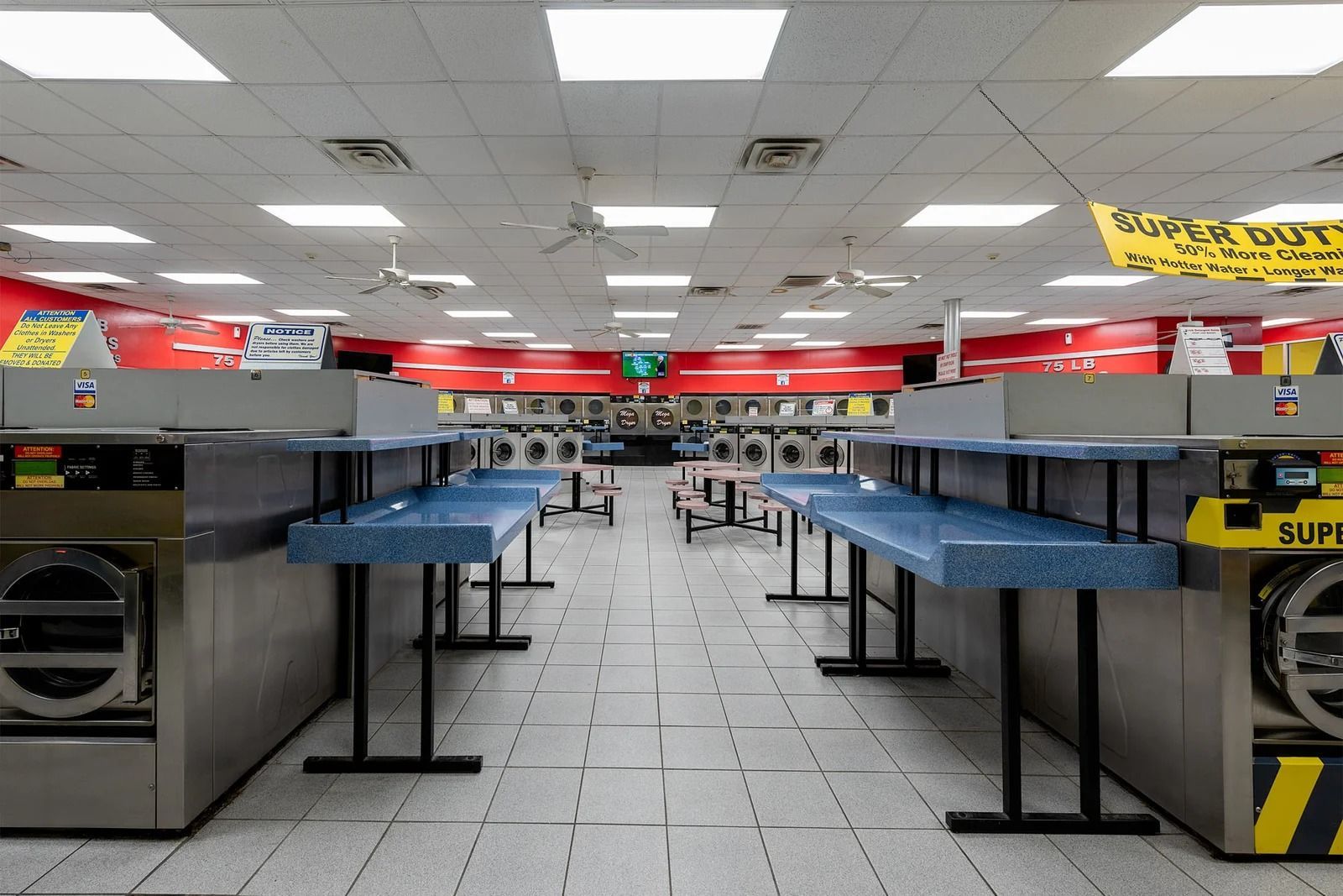 Laundromat interior with rows of washing machines and folding tables, fluorescent lighting, and red and yellow accents.