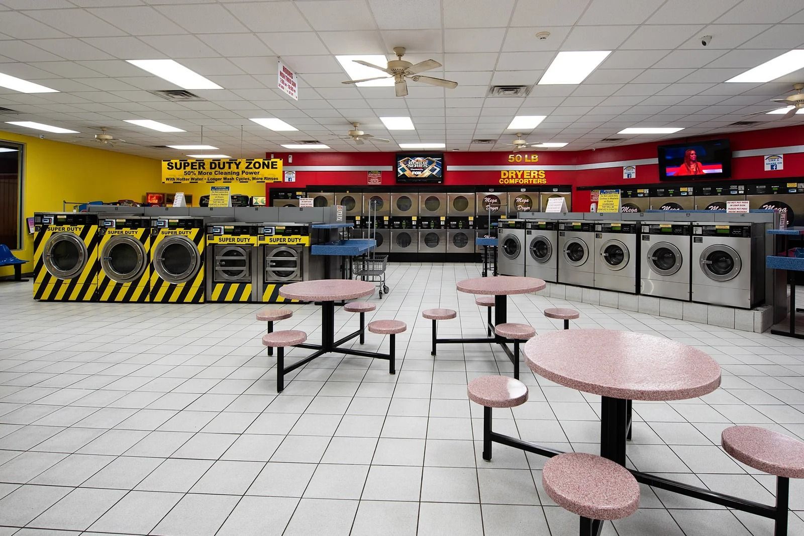 Interior of a laundromat with rows of washers and dryers, tables, and seating. Yellow, red, and white color scheme.