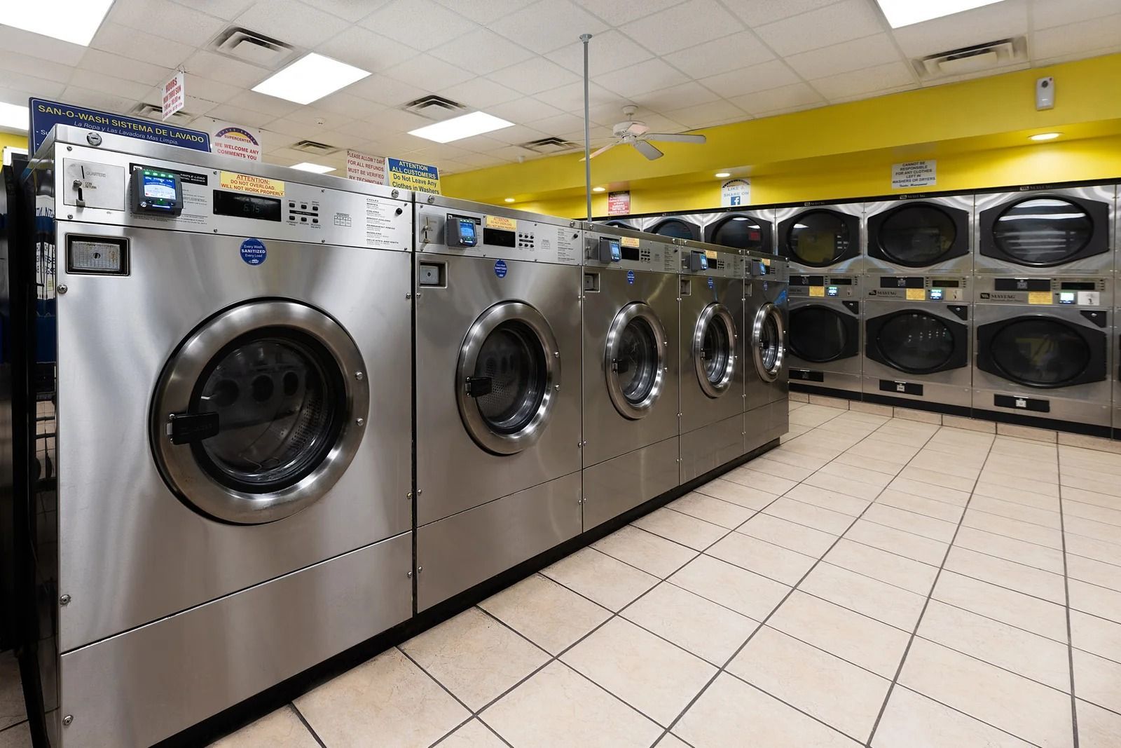 Row of stainless steel washing machines in a brightly lit laundromat with tiled floor and yellow accents.