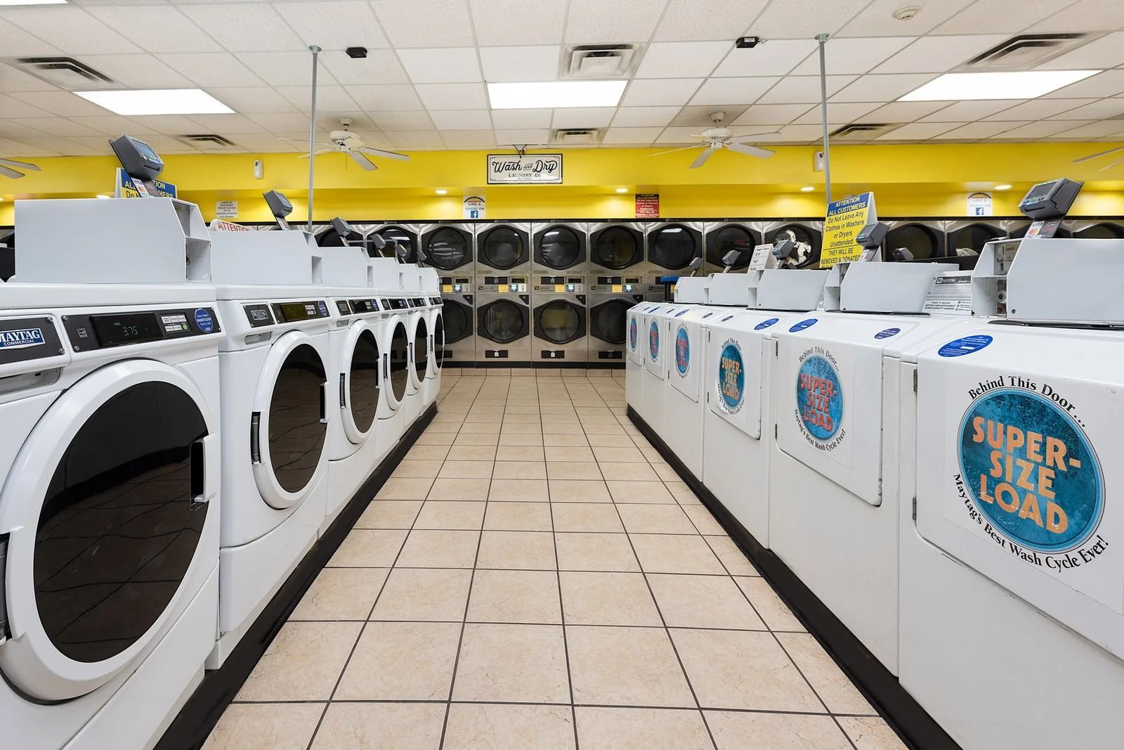 Row of washing machines in a laundromat with a center aisle, yellow walls, and fluorescent lights.