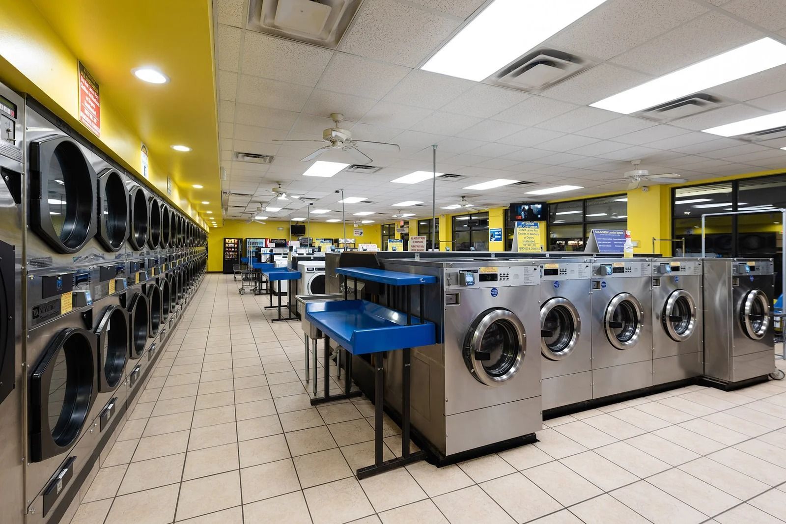 A brightly lit laundromat with rows of washers and dryers, blue folding tables, and yellow walls.