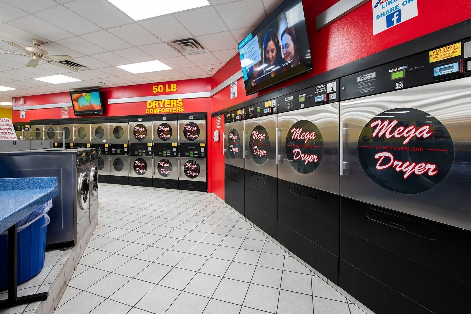 Interior of a laundromat with rows of washers and dryers, a blue counter, and TVs playing. Red and black accents.