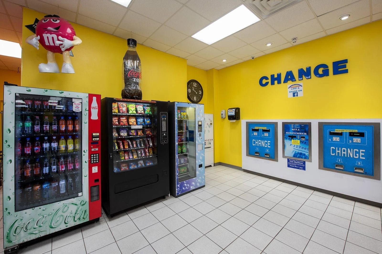 Vending machines and change machines in a brightly lit room with a large M&M figure on the wall.