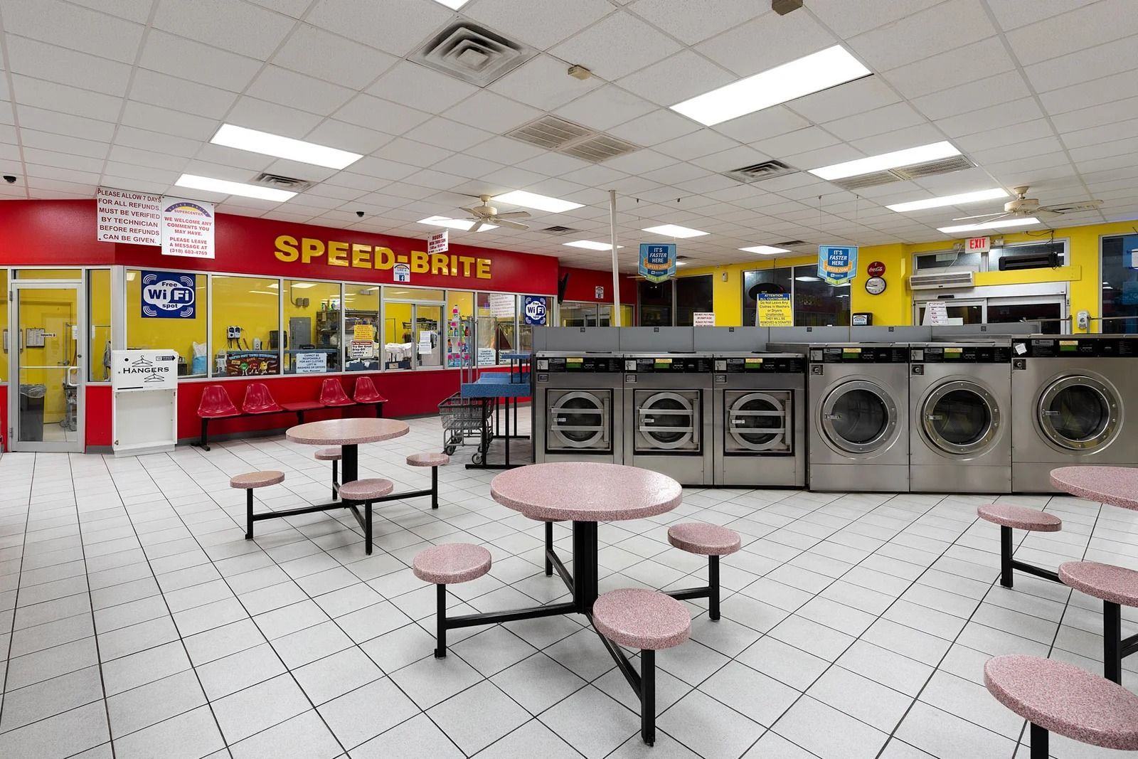 Interior of a laundromat, featuring rows of washing machines, round tables, seating, and a bright red and yellow color scheme.