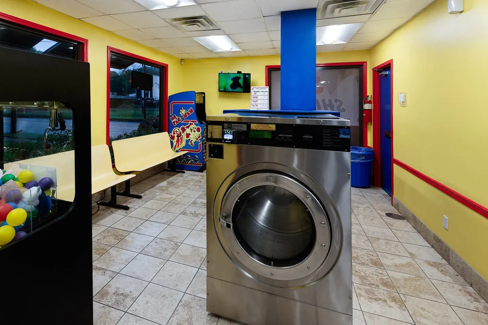 Laundry room with a large washing machine in the center; yellow walls, a bench, and a gumball machine are visible.