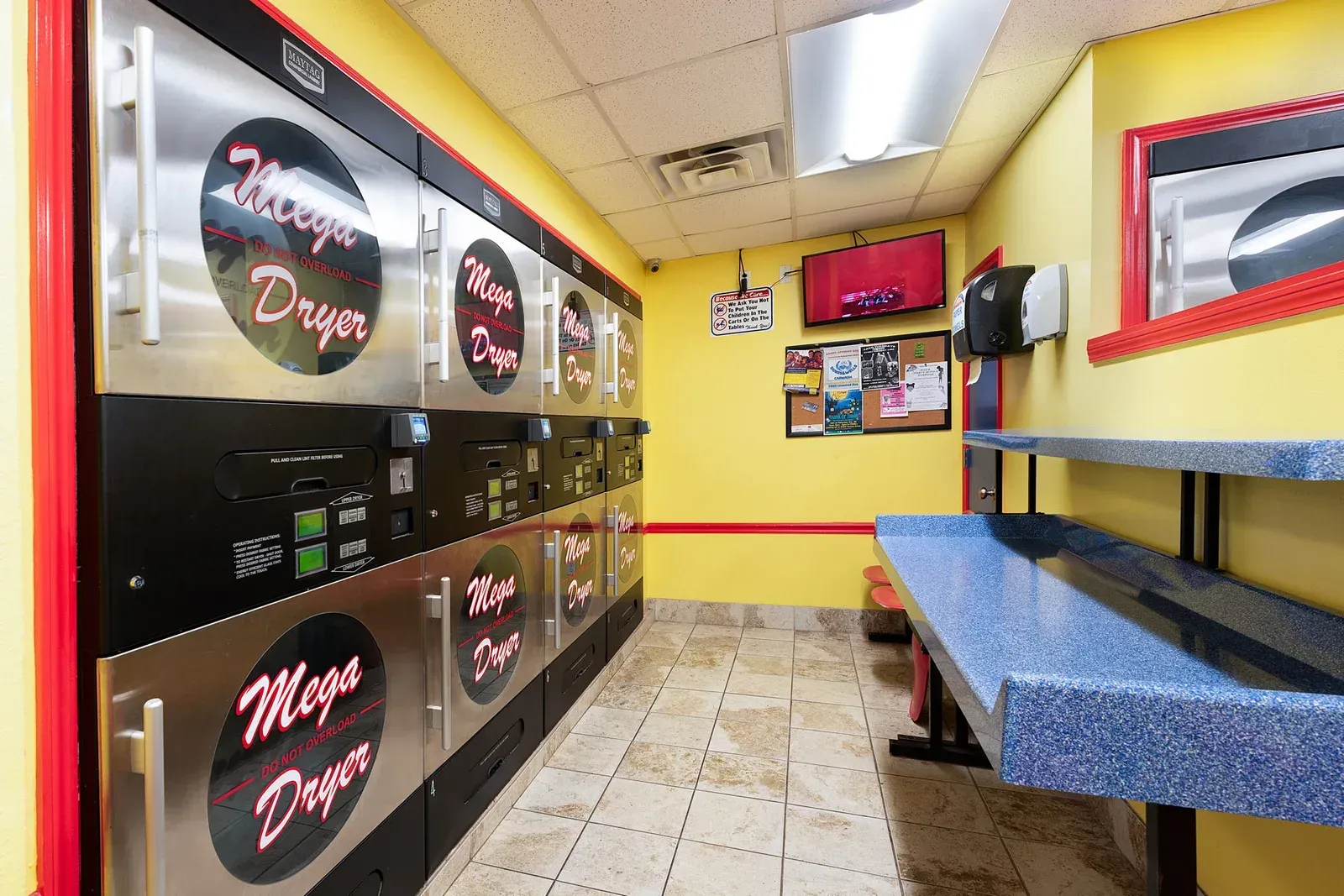Laundromat interior with dryers, folding table, yellow walls, and a television.