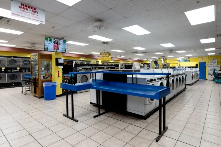 Interior of a laundromat with rows of washers and dryers, blue folding tables, and bright overhead lights.