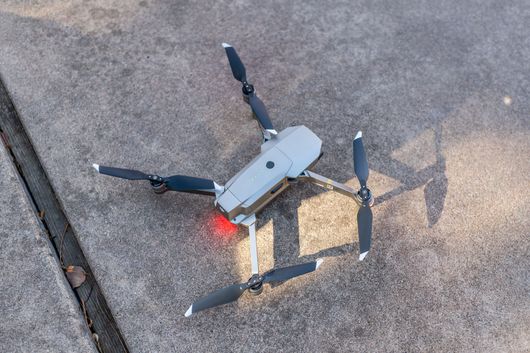 Small gray quadcopter drone hovering low over a concrete surface, with red lights glowing beneath it.