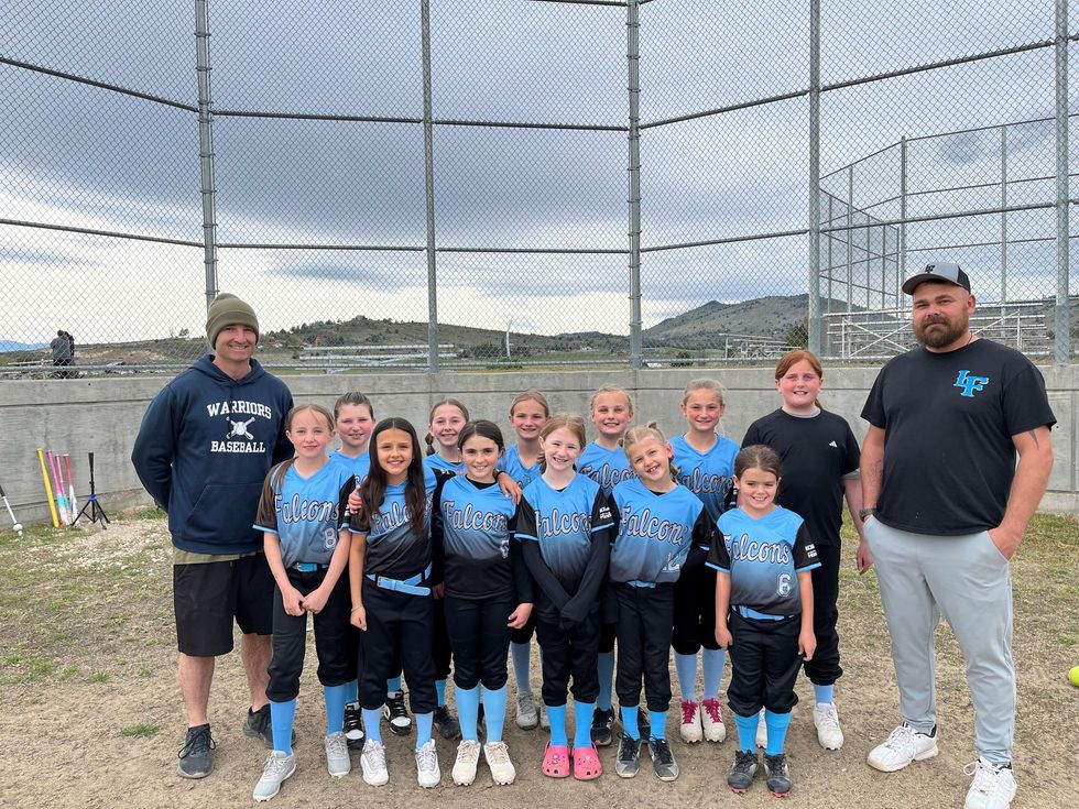 Youth baseball team and coaches posing on a dirt field in front of a chain-link backstop and lake.