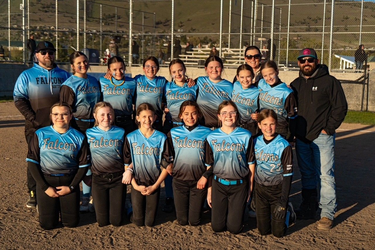Softball team posing outdoors in blue jerseys with a coach beside them on a field