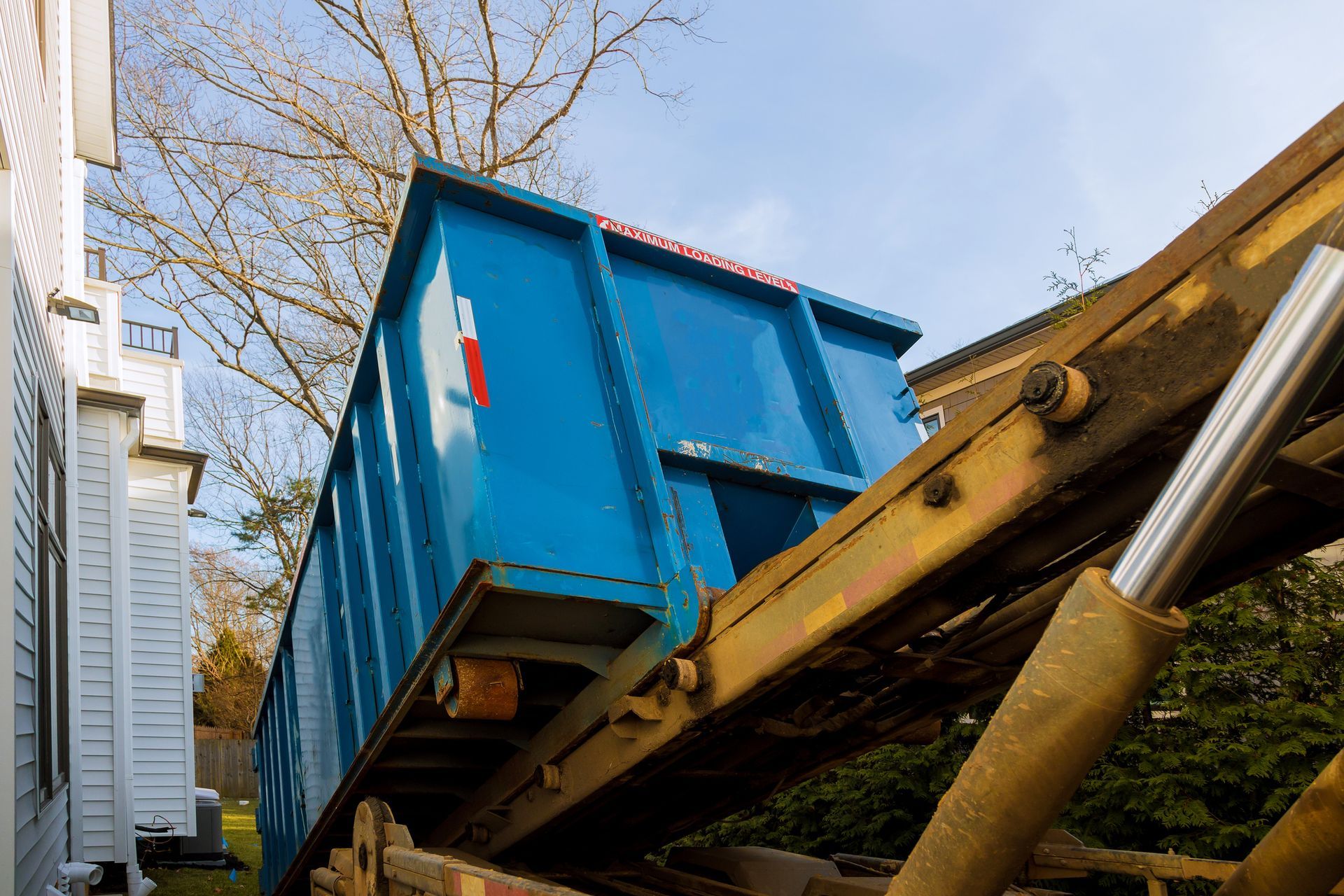 A blue dumpster is being loaded onto a truck in front of a house.