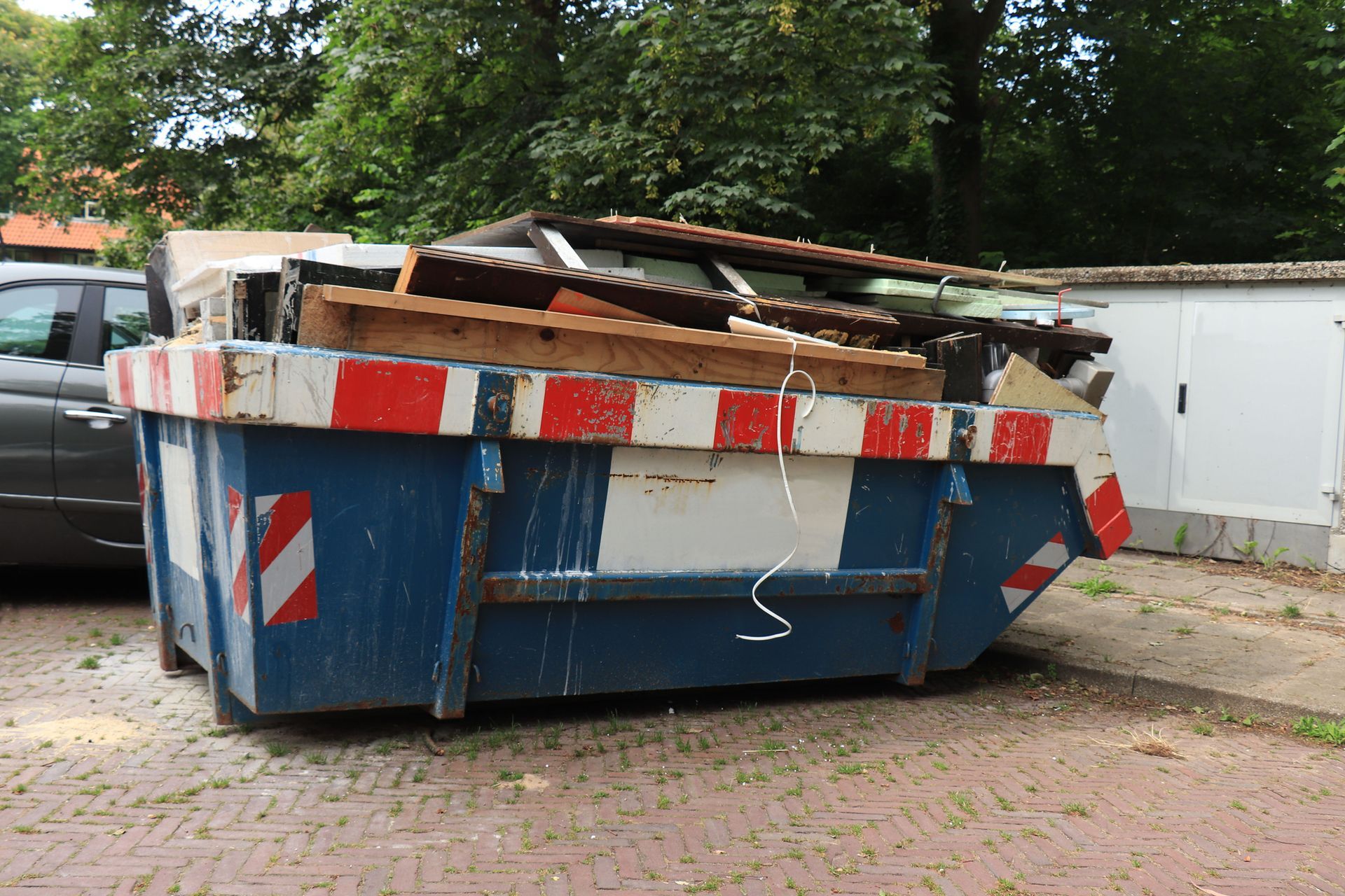 A blue dumpster filled with junk is parked next to a car.