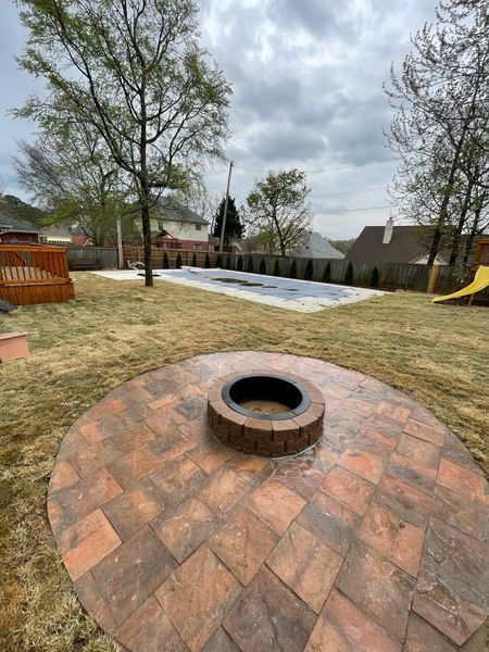 Backyard with a brick fire pit, swimming pool, and trees under a cloudy sky.