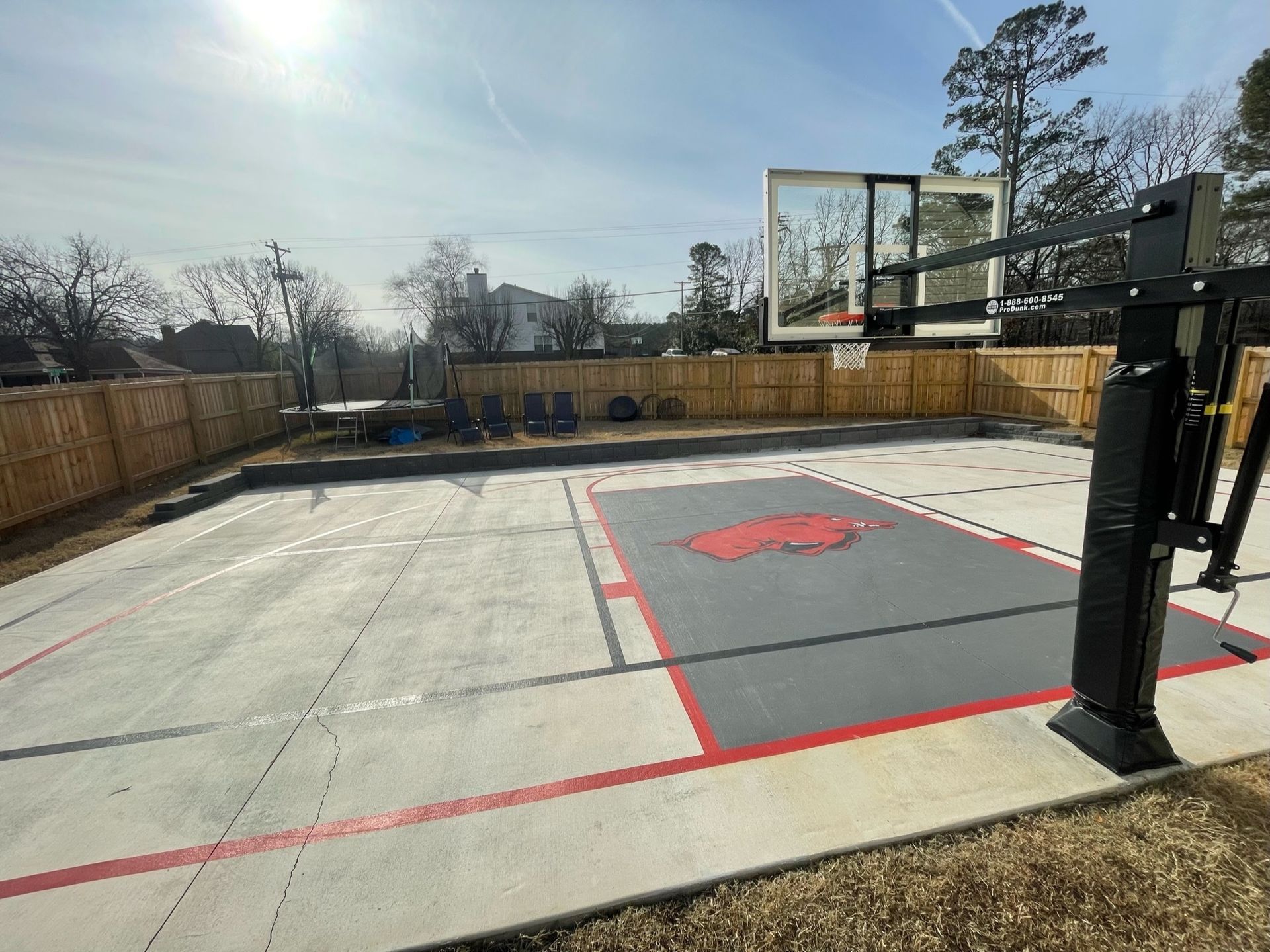 Outdoor basketball court with red, white, and gray lines, black hoop, and red logo. Wooden fence surrounds the court.