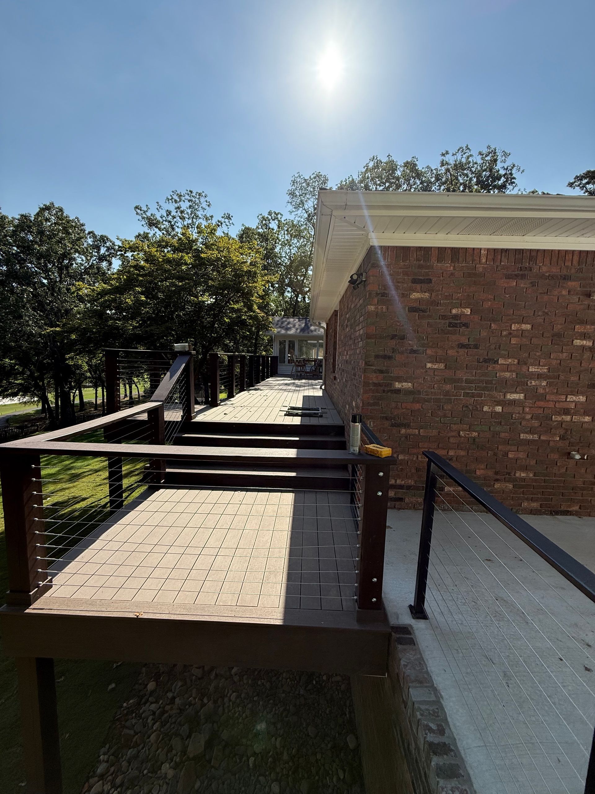 Wooden deck next to a brick house. Sunny day with trees in the background.