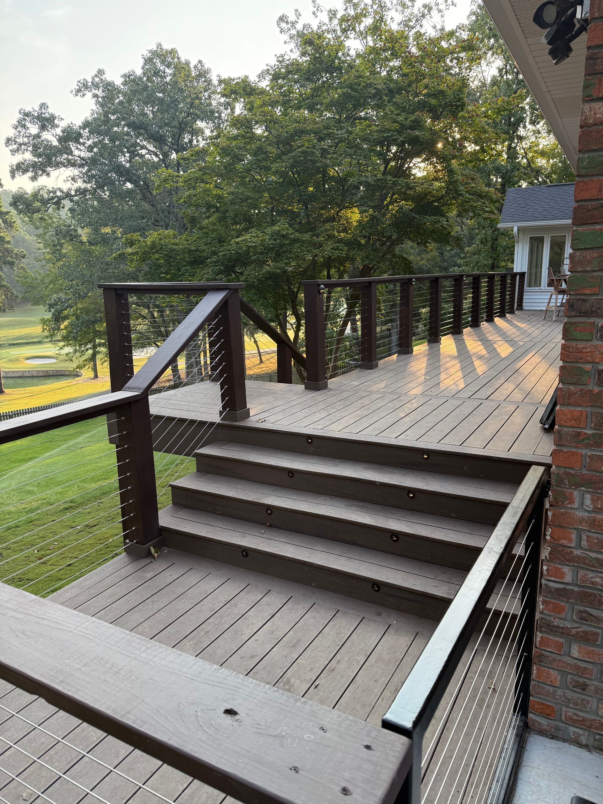 Wooden deck with steps and dark brown railings; cable infill, brick wall, and green lawn in the background.