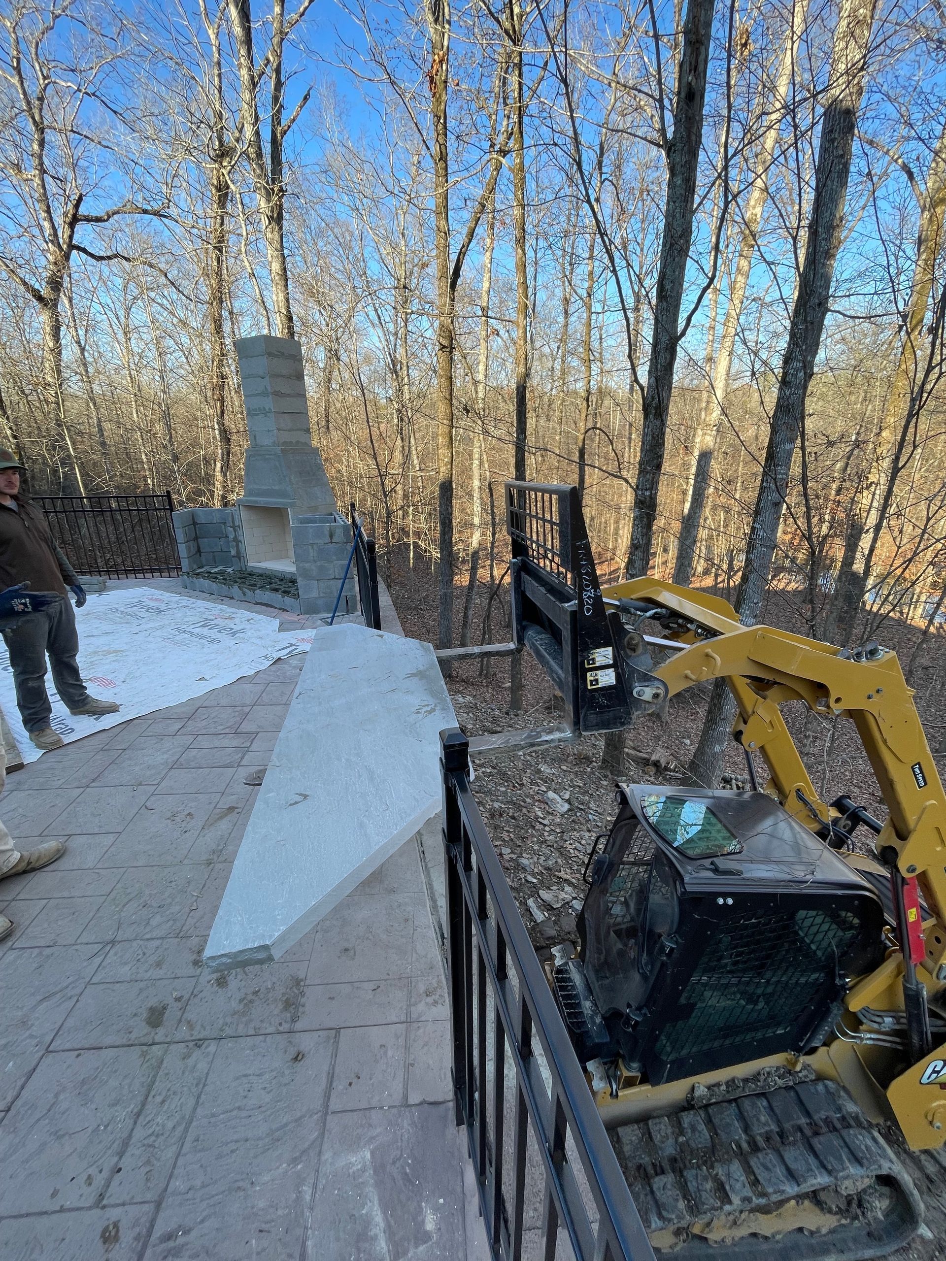 A yellow excavator on a patio removes dirt. A man stands nearby. Trees and an outdoor fireplace are in the background.