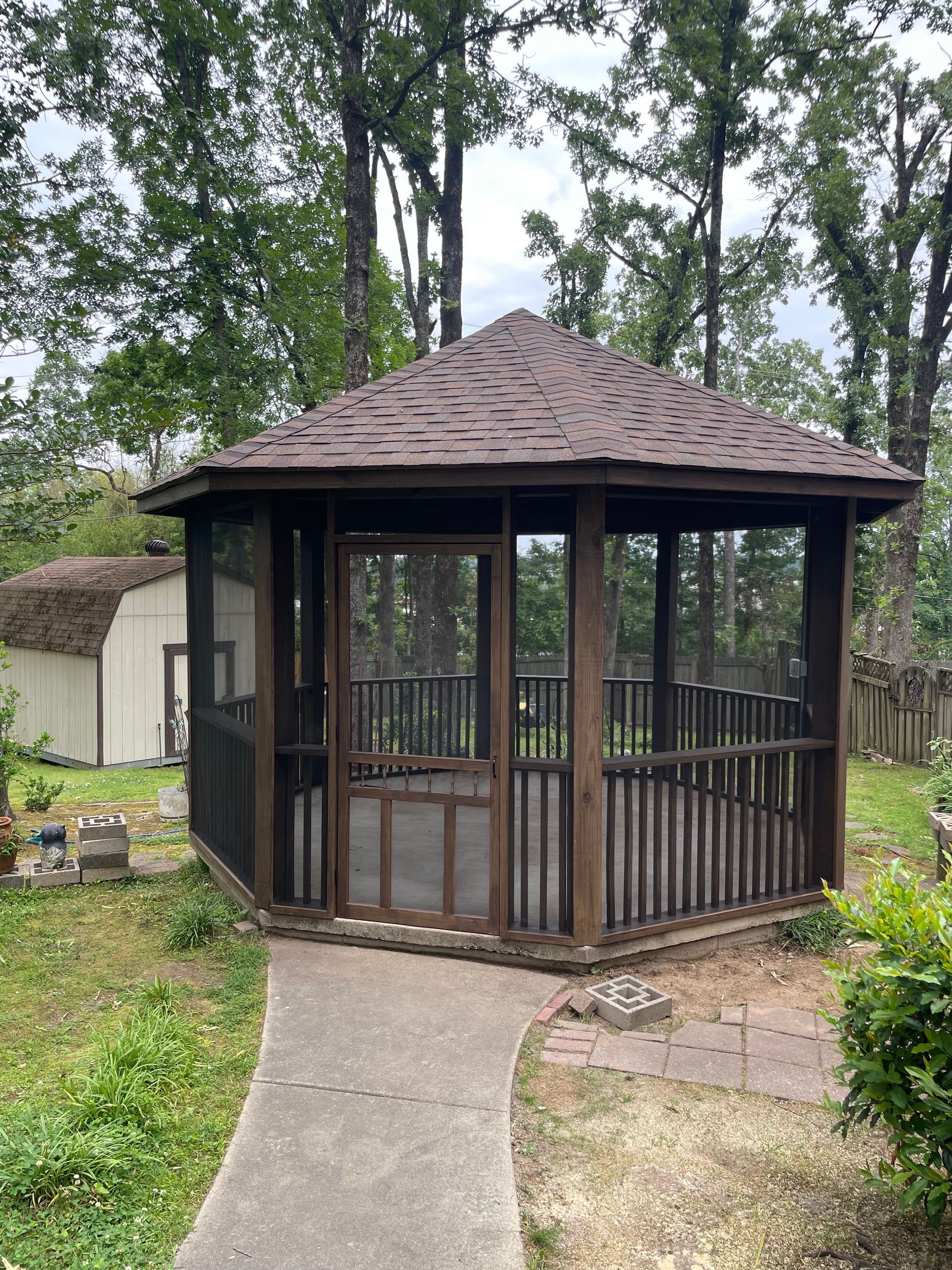 Screened gazebo with a brown roof and wooden railings in a backyard. Concrete path leading to it.