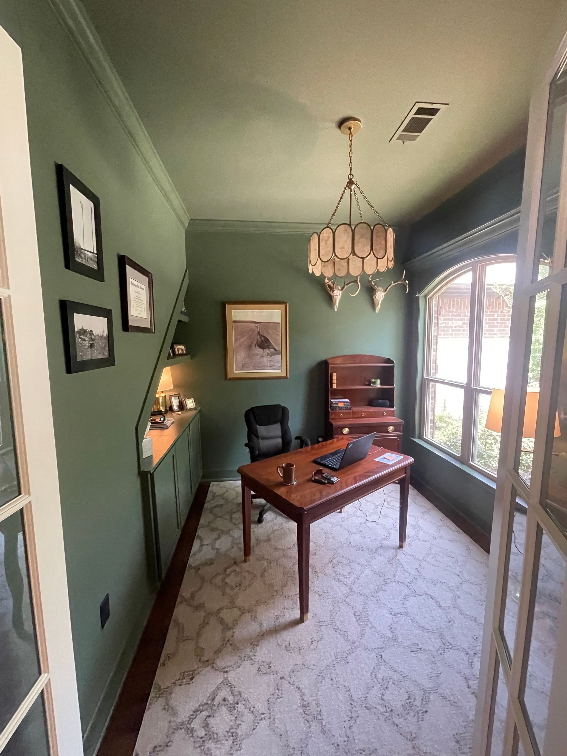 Office with green walls, a wooden desk, bookshelf, and framed artwork. A chandelier hangs from the ceiling.