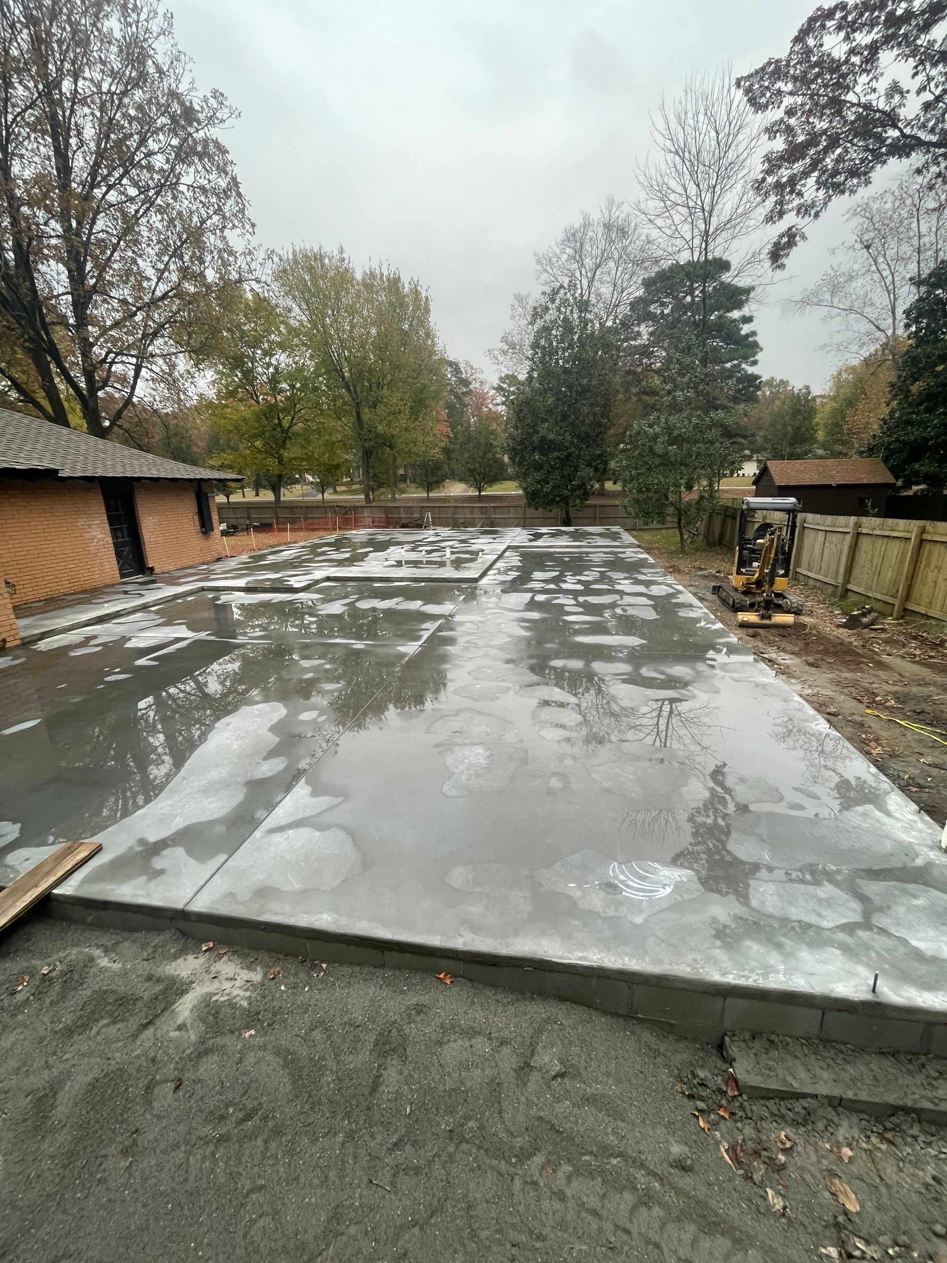 Newly poured concrete foundation, reflecting overcast sky. Brick building and trees in background.