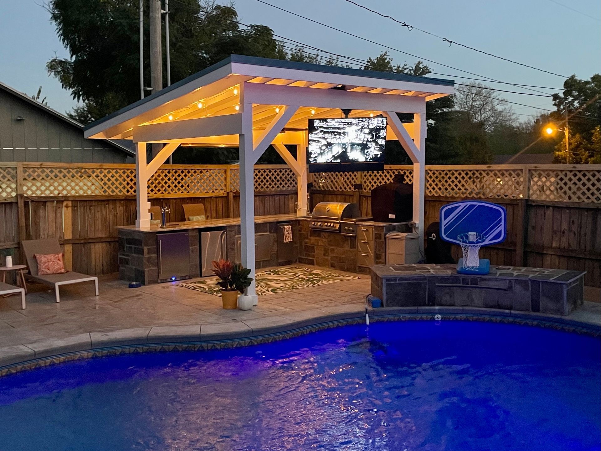Outdoor kitchen with a pool. A covered structure features a TV, grill, and seating area. Lit by fairy lights.
