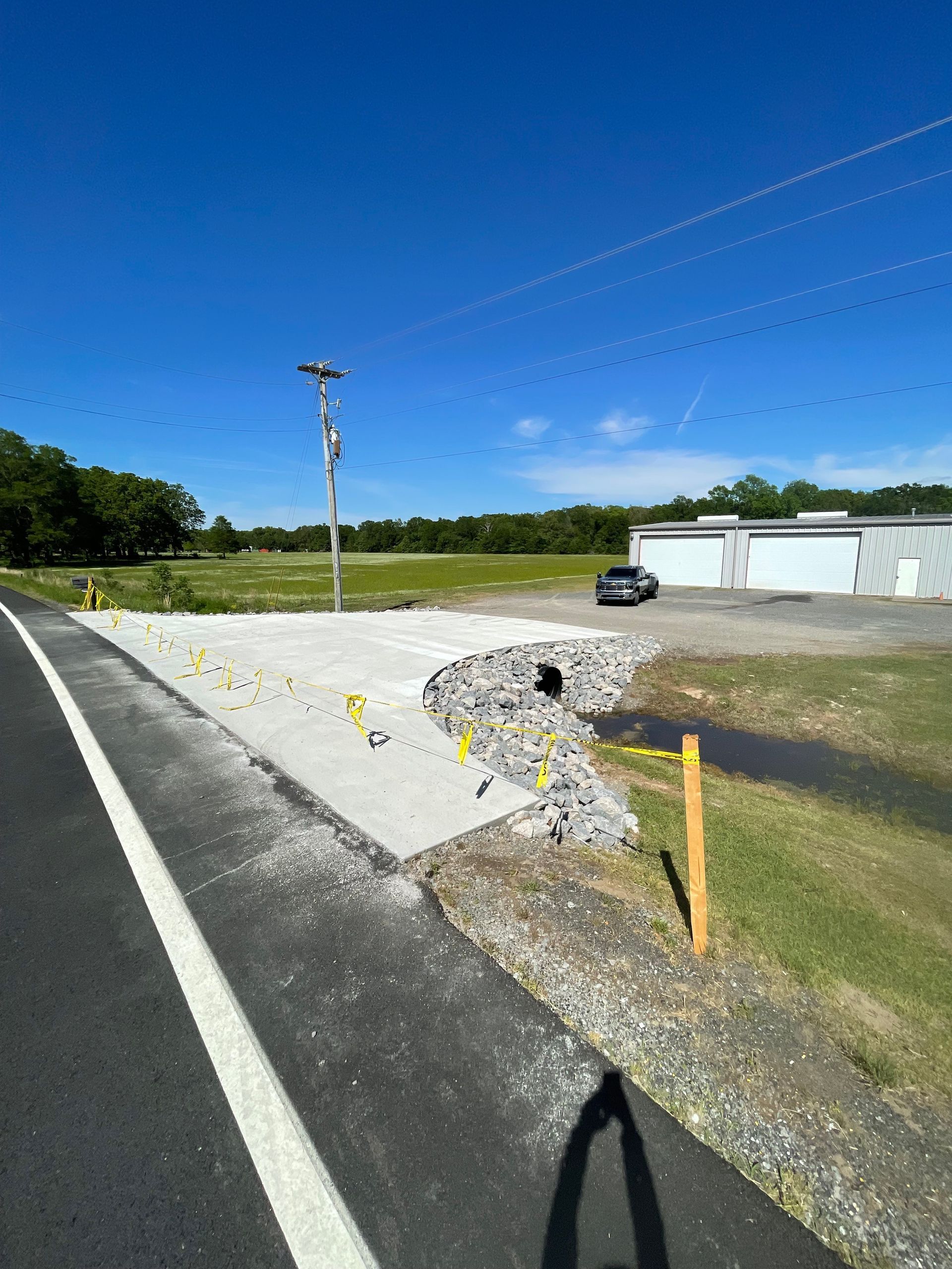 A damaged roadside with exposed concrete and a drainage pipe next to a paved road and a building under a blue sky.