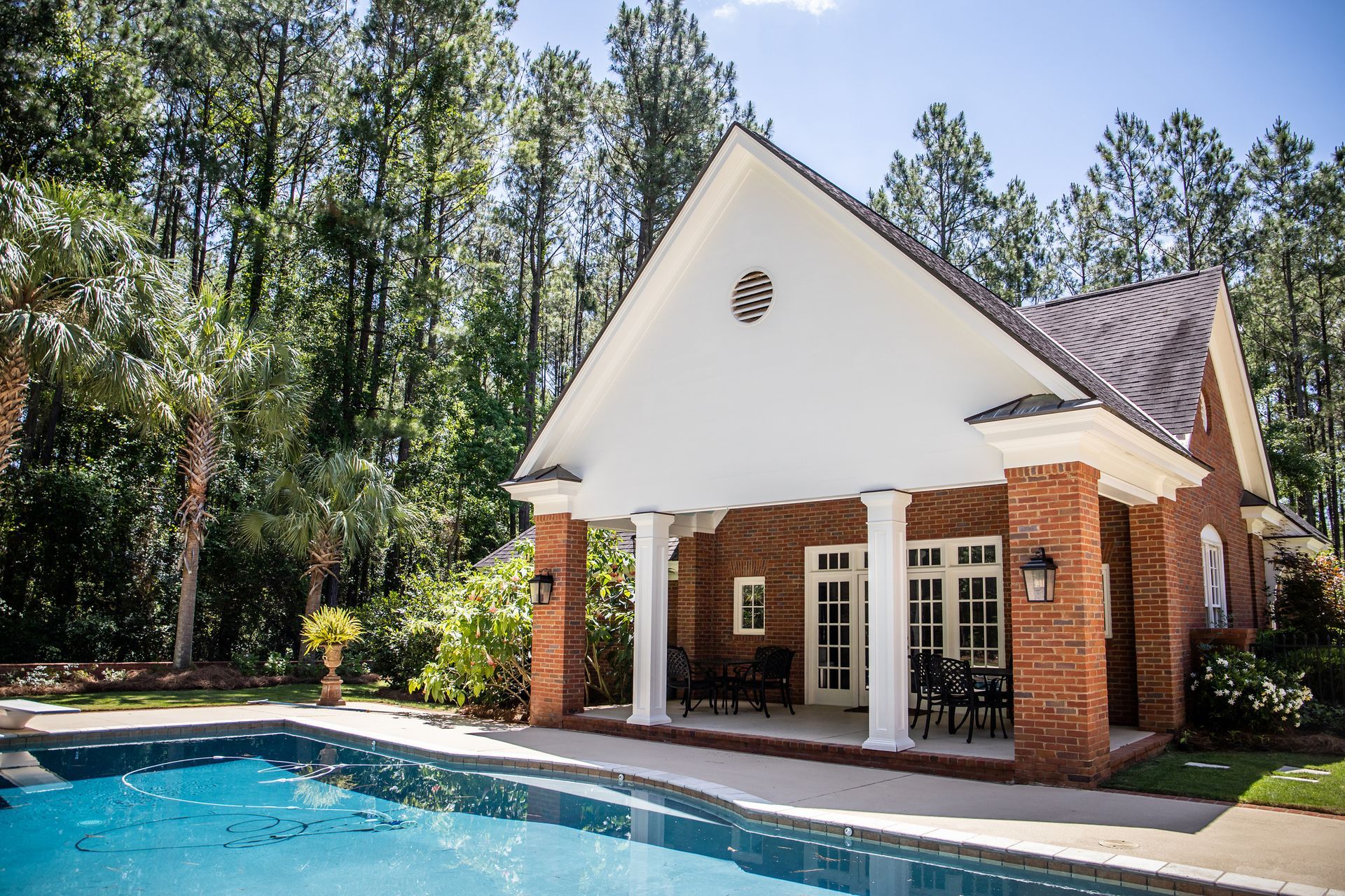 Poolside brick building with white pillars and roof. Sunny day with trees and pool.