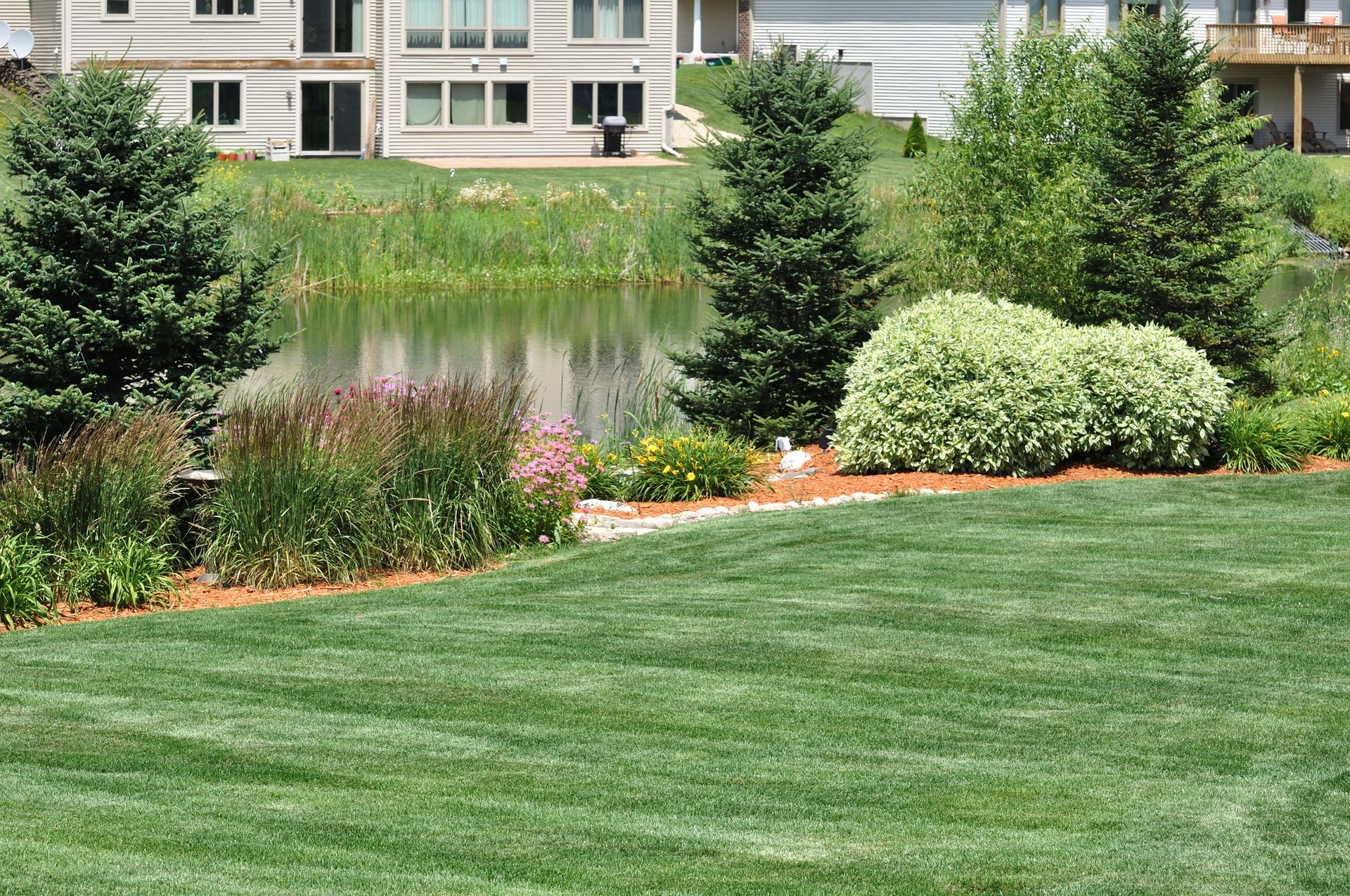 Lush green lawn borders a landscaped pond with trees and flowers, with a two-story house in the background.