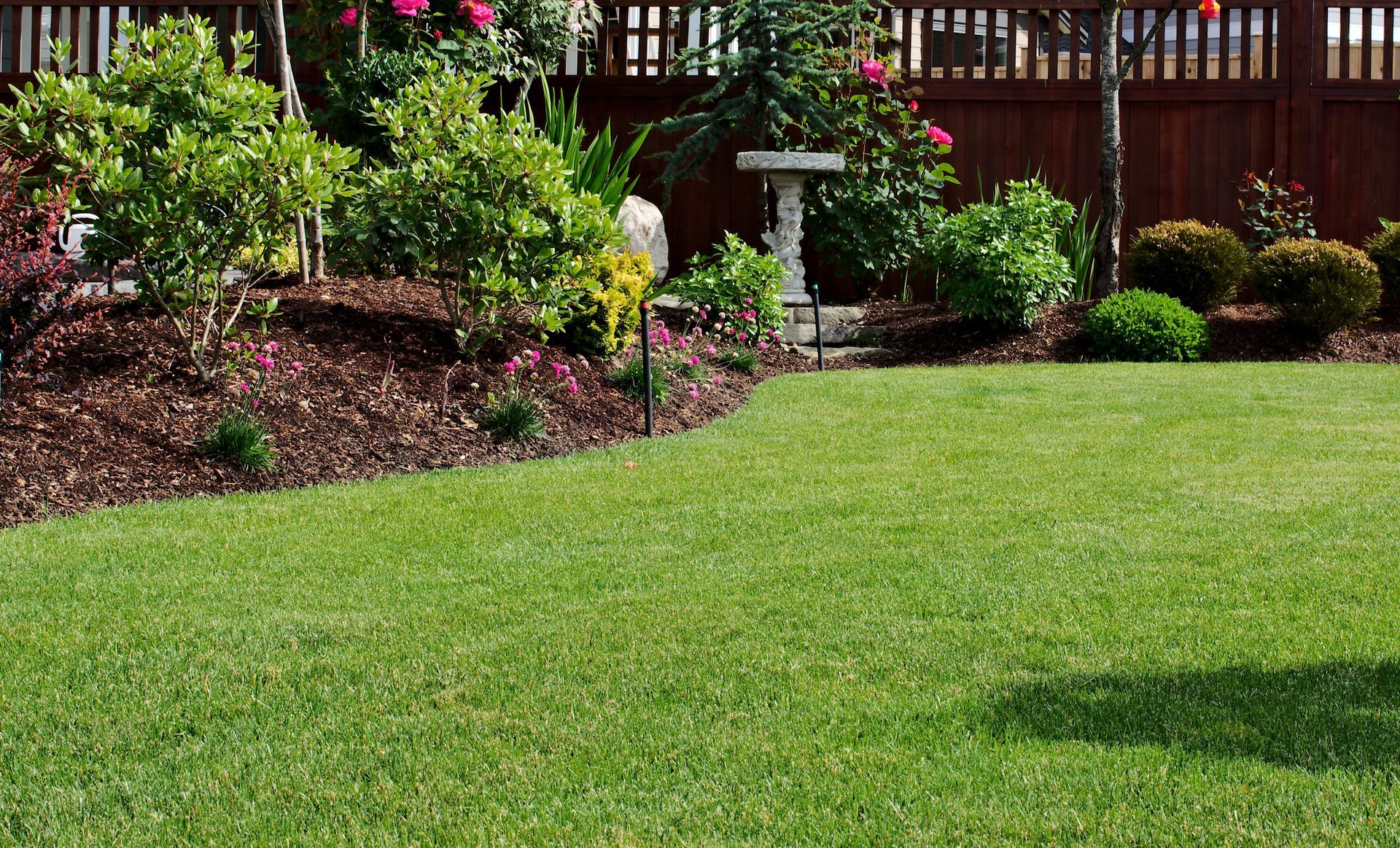 Lush green lawn borders a garden bed with blooming flowers and shrubs, wooden fence in background.