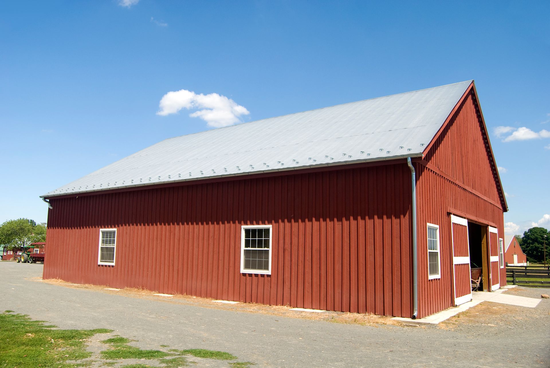 Red barn with a silver roof against a blue sky. Gravel ground and small windows.