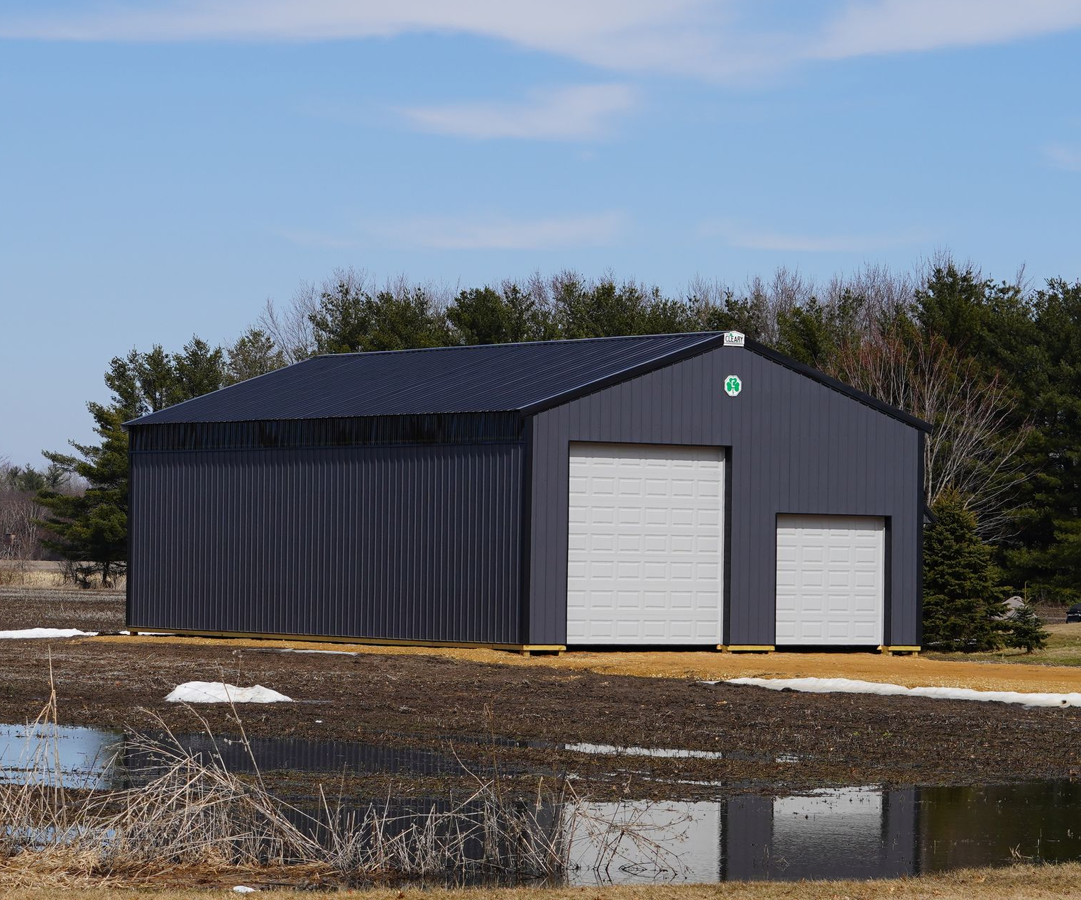 Gray metal building with a dark roof and two white garage doors, near a pond with some snow.