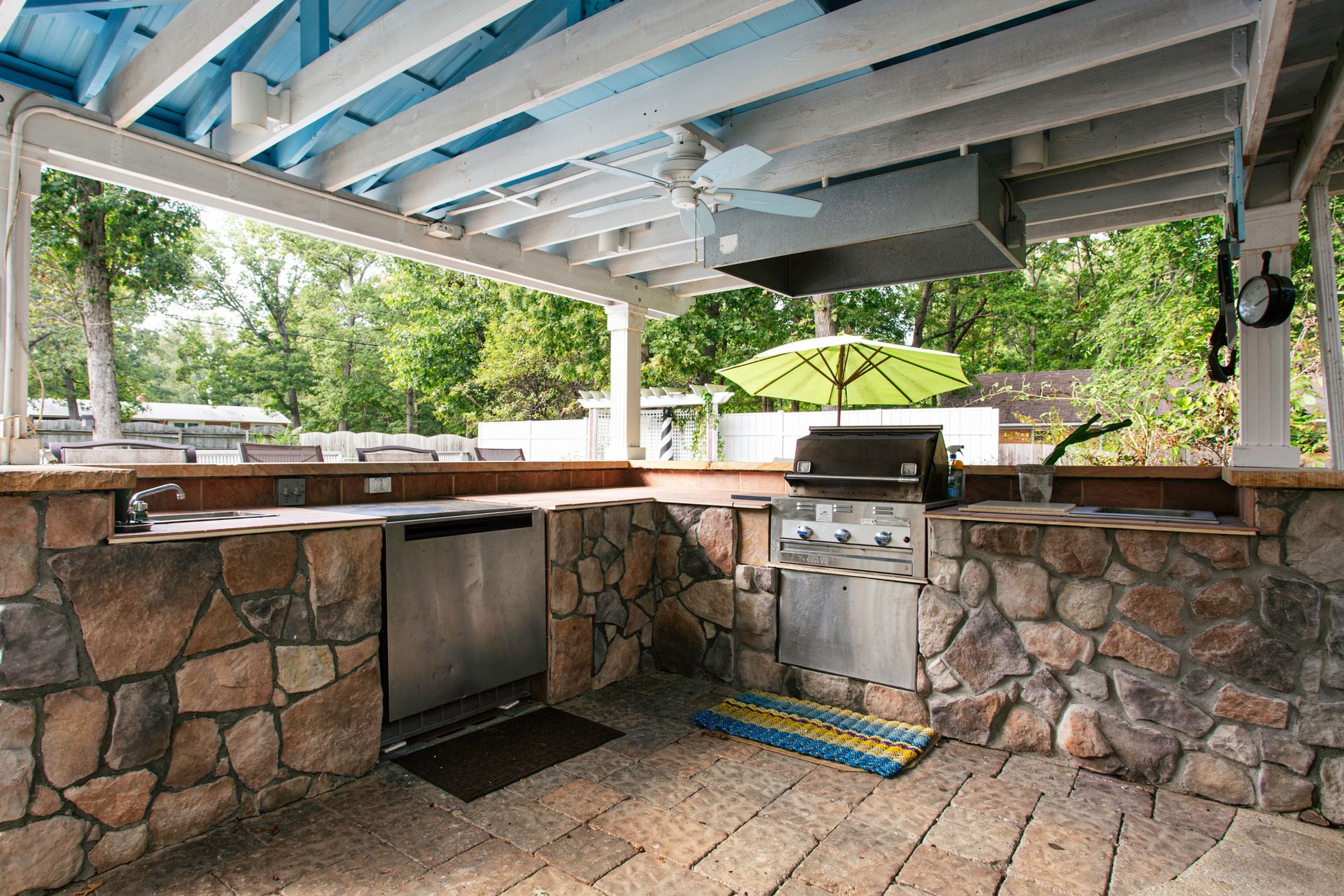 Outdoor kitchen with stone counters, grill, and overhead shelter; green trees in background.