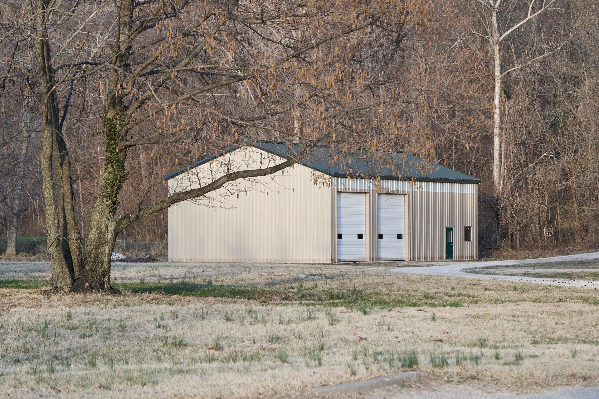 Light-colored building with two garage doors sits near bare trees, on a patch of frosted grass, in a wooded area.