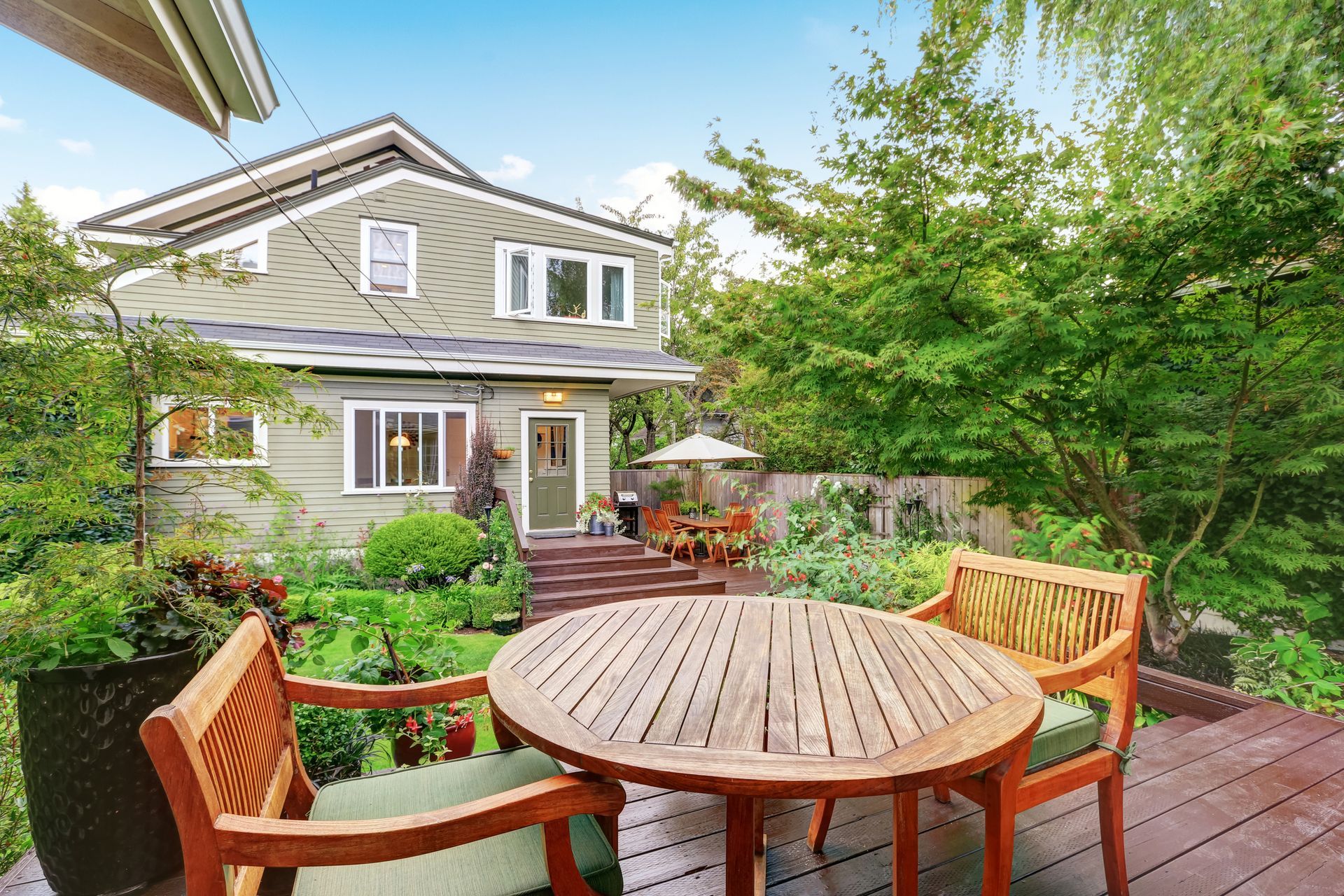 Wooden patio furniture on a deck, with a two-story green house in the background. Lush garden setting.