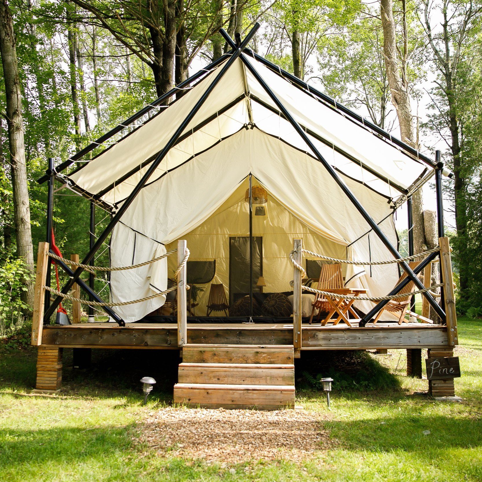 Canvas tent on a wooden platform deck, in a wooded area. The tent has a black frame, porch, and steps.