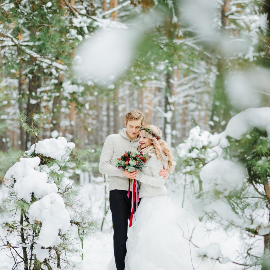 Couple embraces in snowy forest; woman in wedding dress, man in sweater, holding bouquet.
