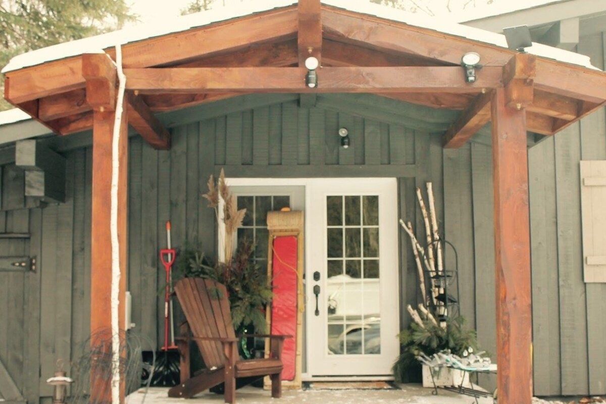 Wooden porch over white double doors of a gray cabin, decorated with greenery and an Adirondack chair.