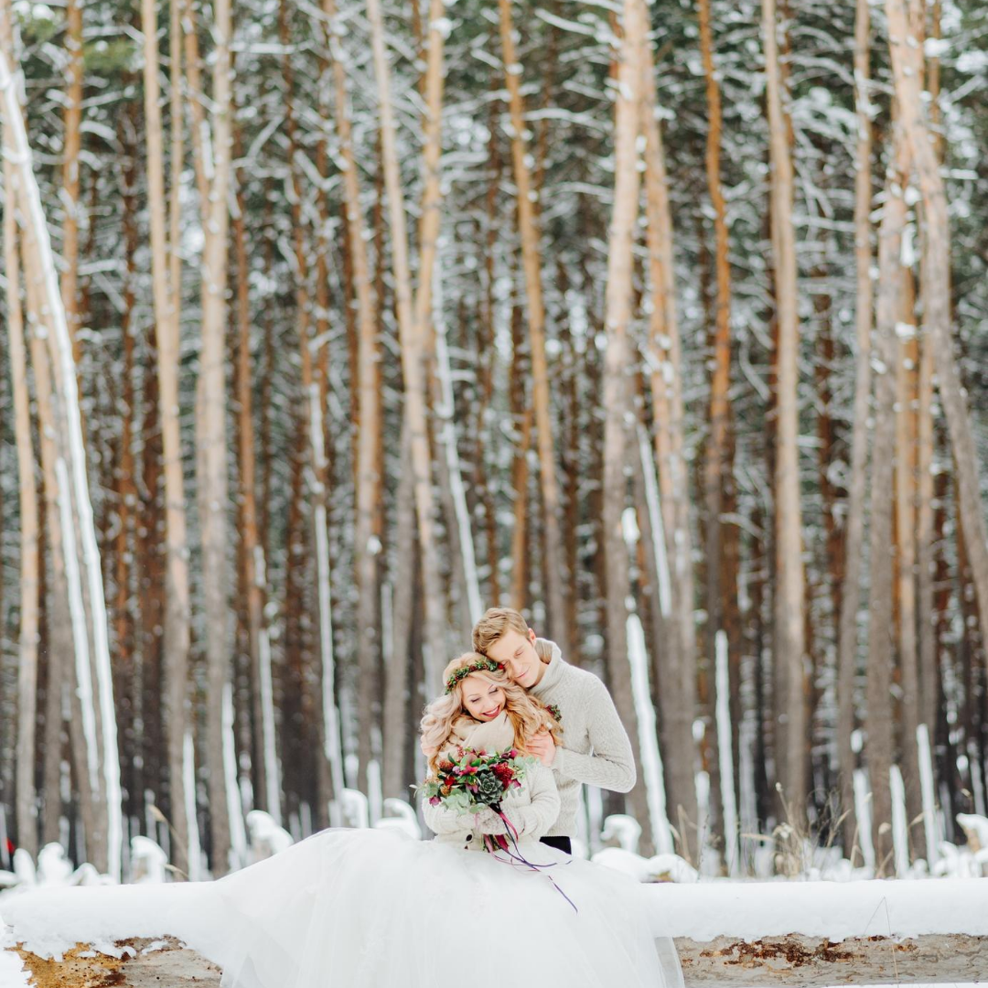 Couple embraces in a snowy forest. Woman in a white dress, man in a sweater, both near a wooden fence.