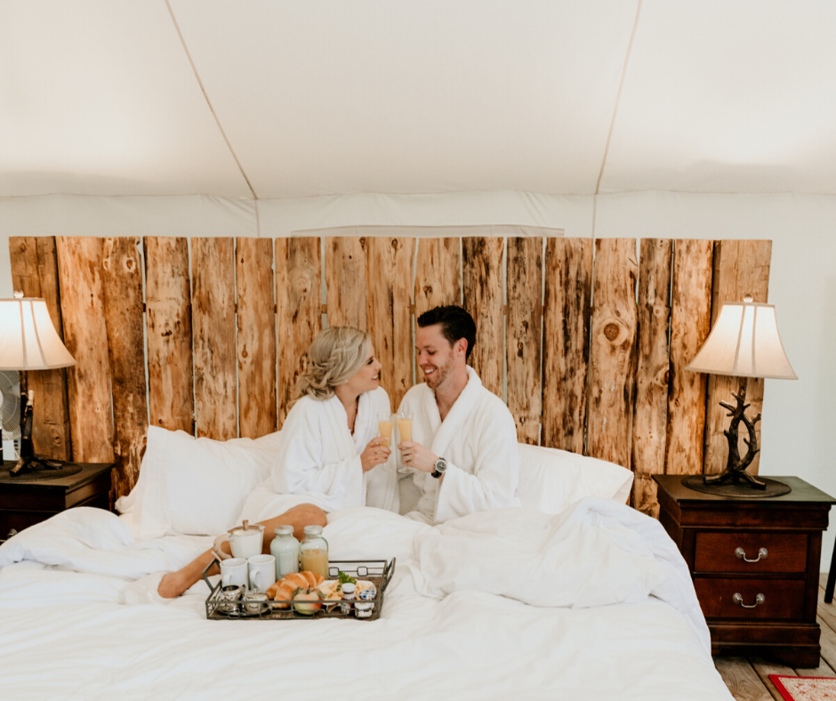 Couple in robes share a toast in bed, breakfast tray, wood headboard, lamp, and bedside tables.