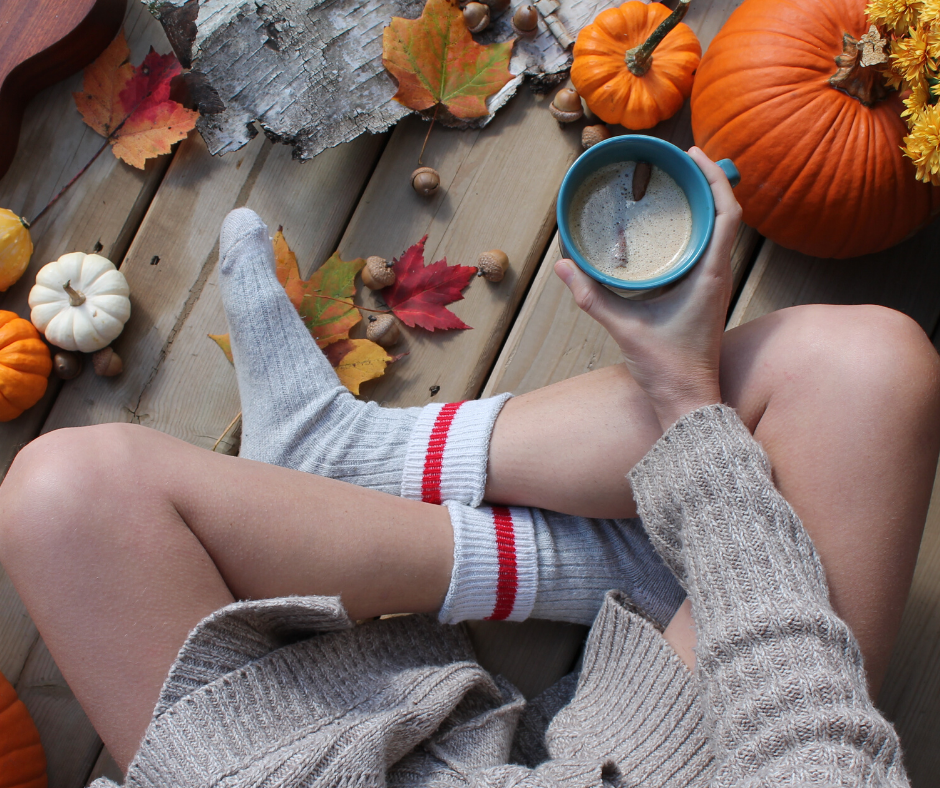 Person holding coffee, resting on wooden deck with pumpkins, autumn leaves, and nuts; wearing cozy sweater and socks.